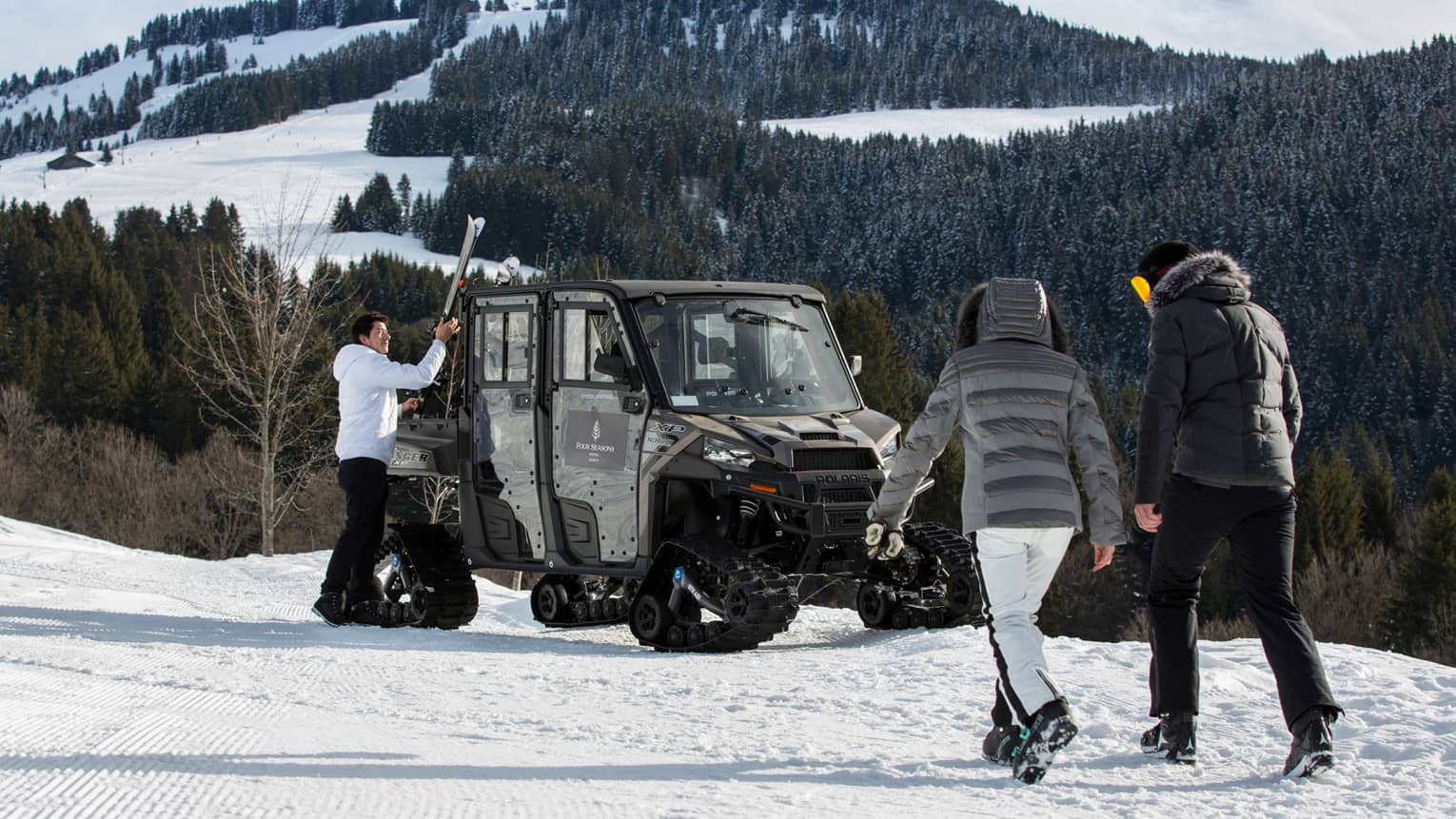 Two people in snowsuits walk toward ski concierge at large snowmobile