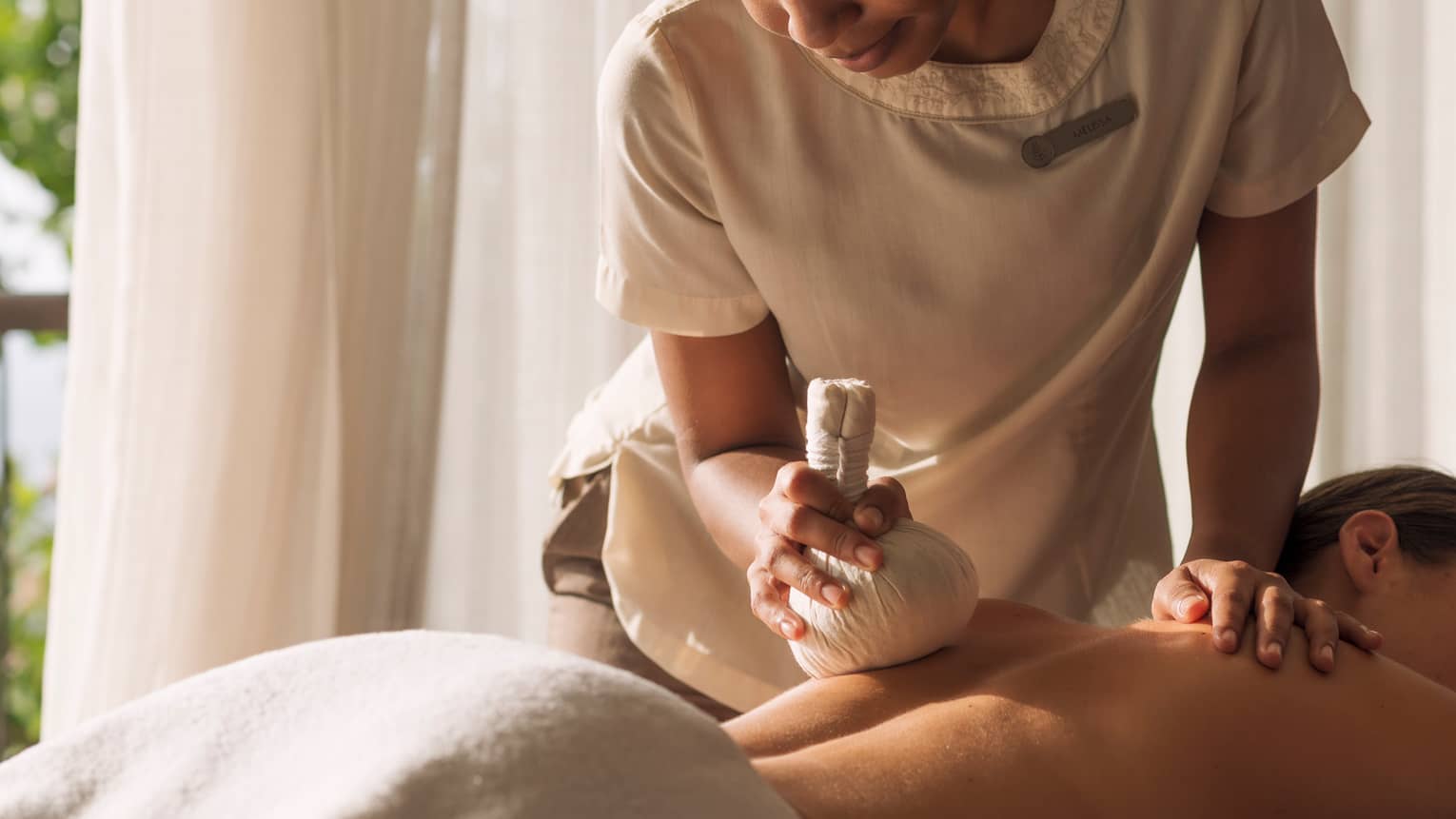 Woman holds stuffed round cloth over woman's bare shoulders as she lies on massage table