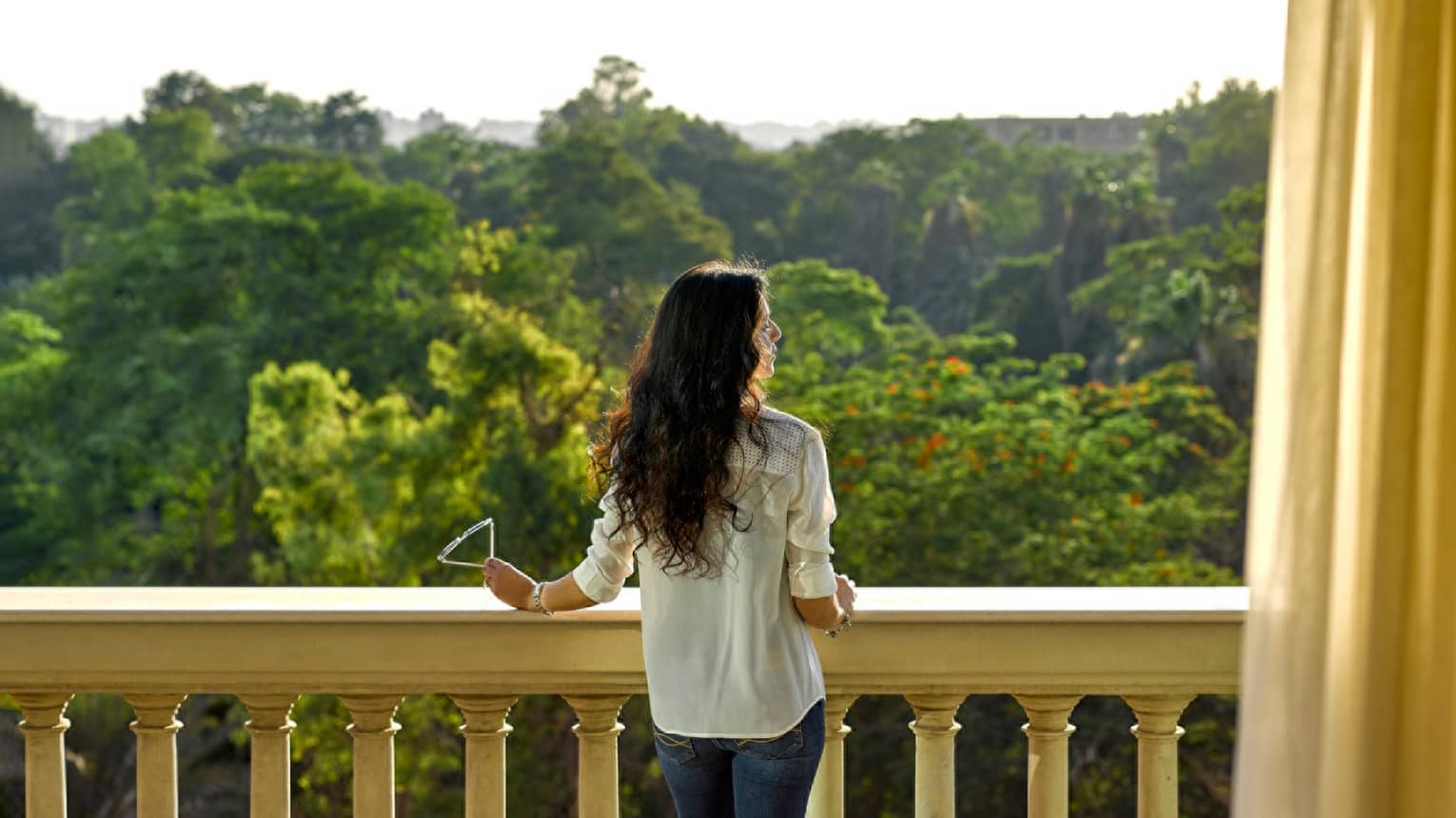 Woman in jeans, button-up shirt looks out over balcony at green zoological gardens canopy