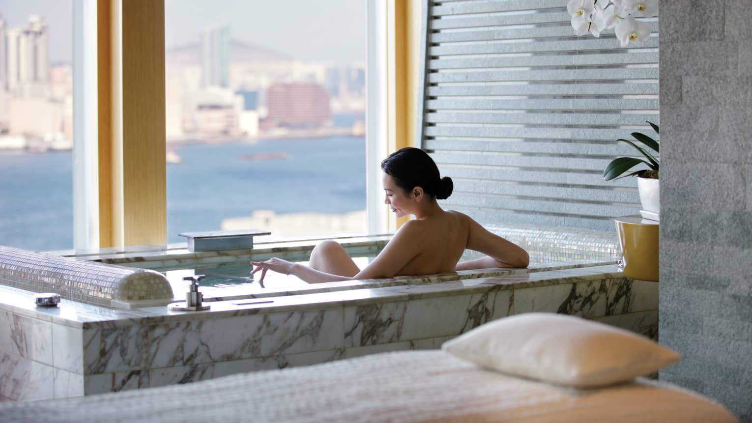 Woman's bare back above marble spa tub in tile bathroom by window with water, city views