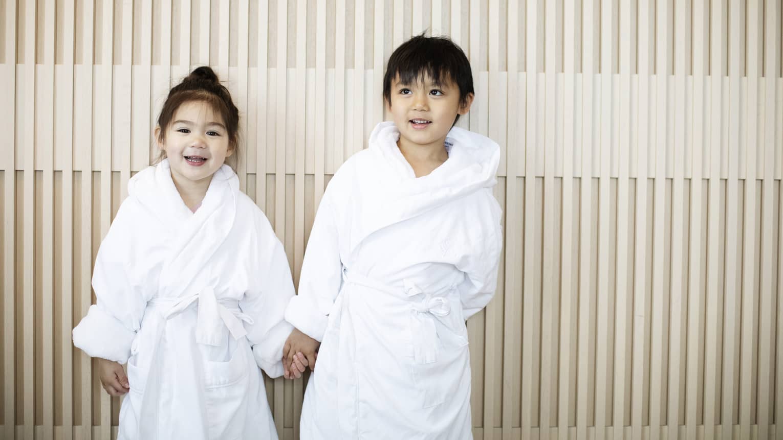 Two children dressed in white bathrobes smile while holding hands