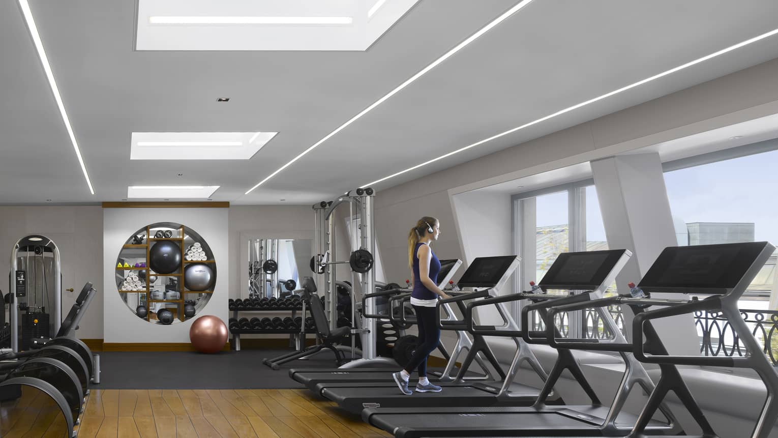 A guest walks on one of a bank of treadmills facing windows in a bright, modern gym with a striking geometric wood floor.
