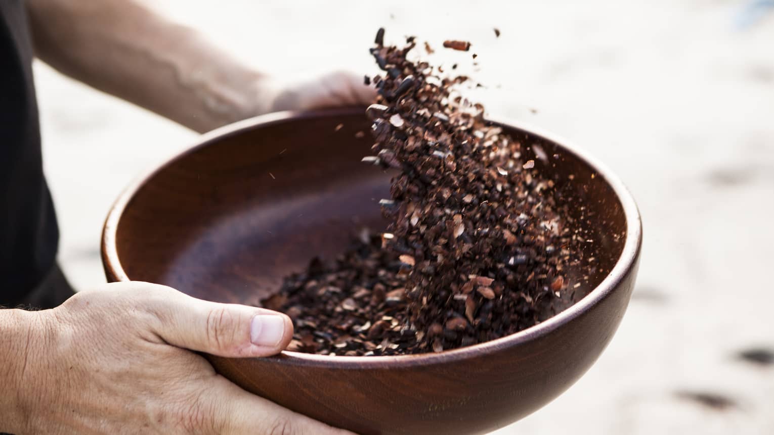 Close-up of hands holding dark wood bowl, shaking crushed cocoa