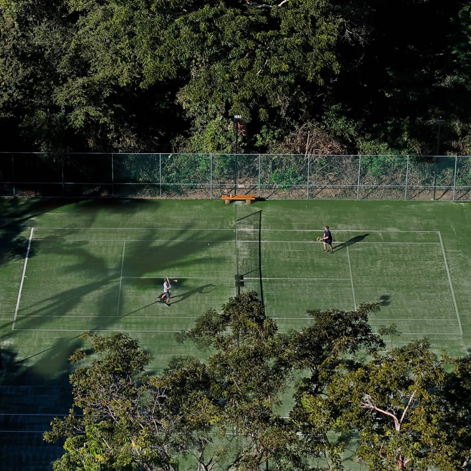 Two people play tennis on a green court surrounded by trees.