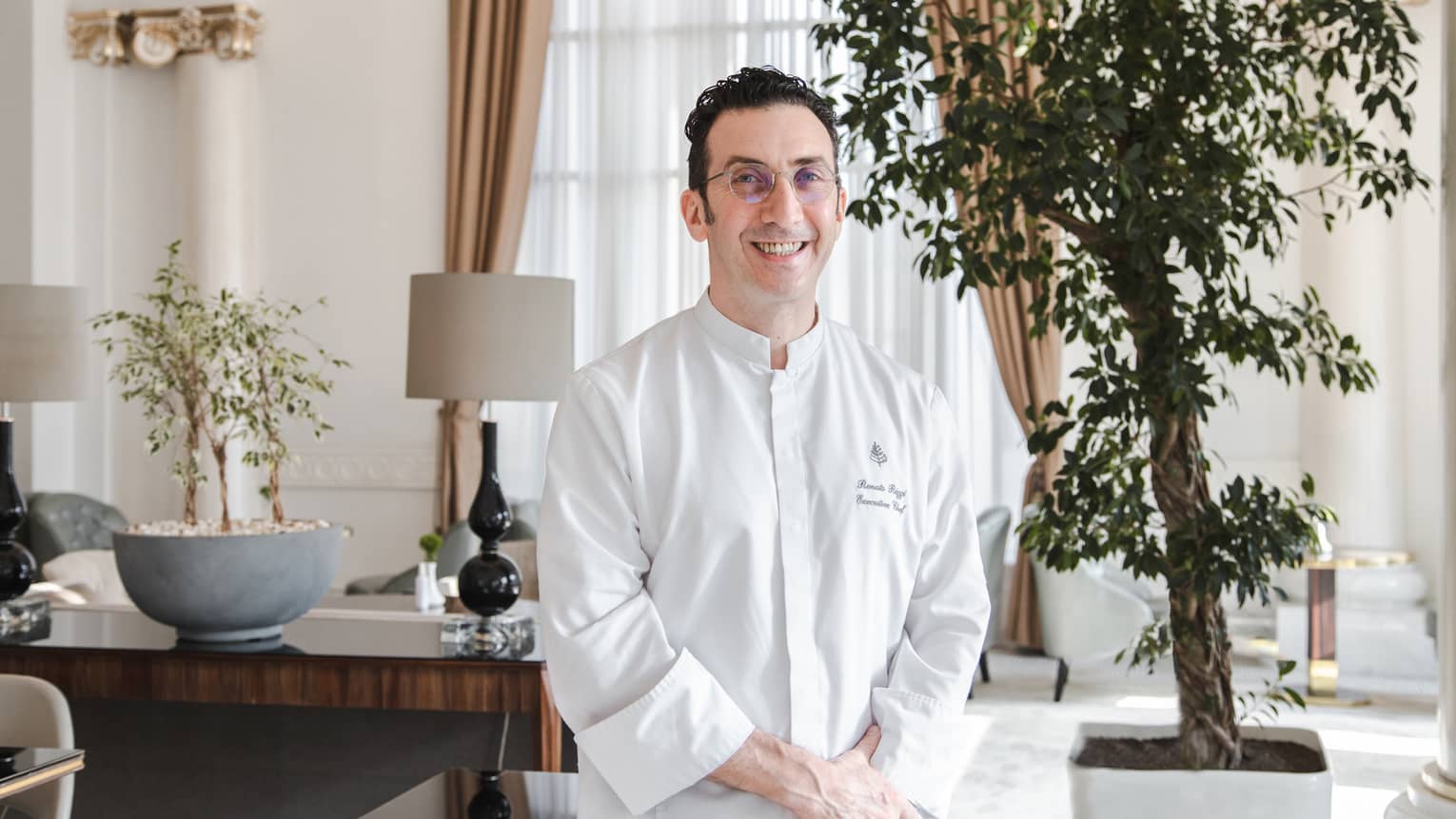A chef in a white chef's coat sitting in a well lit room near a potted plant.