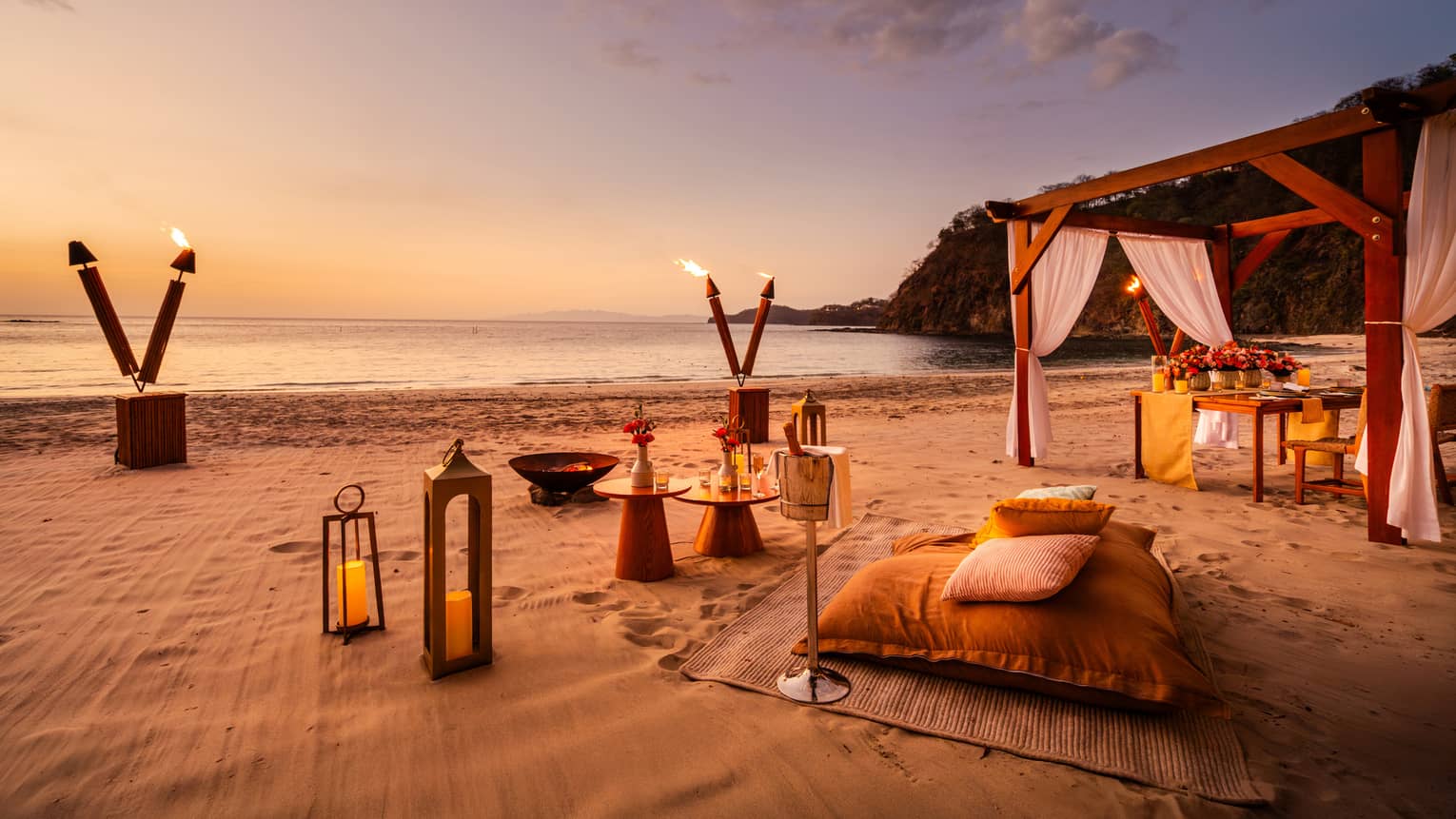 Sunset beach picnic set up with torches, lanterns, large cushions and small round tables
