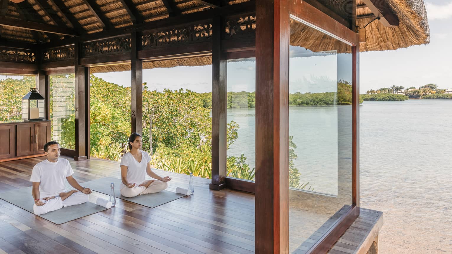 Two people wearing white seated in yoga poses in open-air spa yoga pavilion by beach