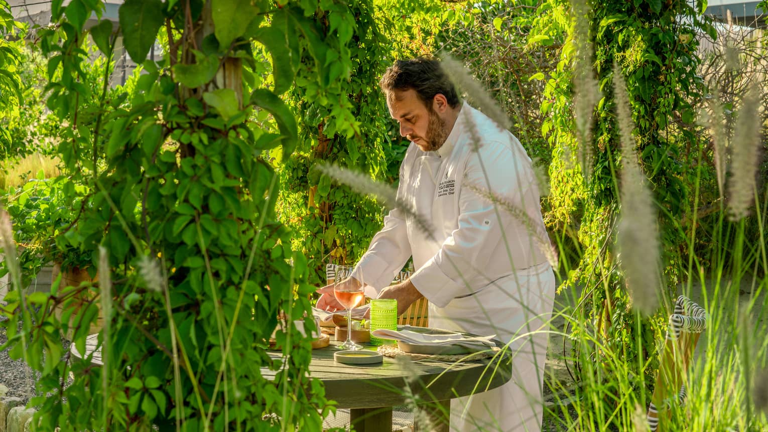 A chef sets food on a table in a lush outdoor dining setting