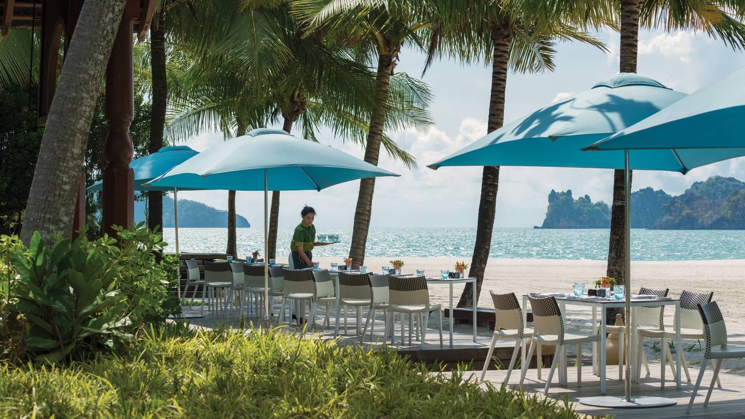 Server sets patio dining tables under blue umbrellas along beach 