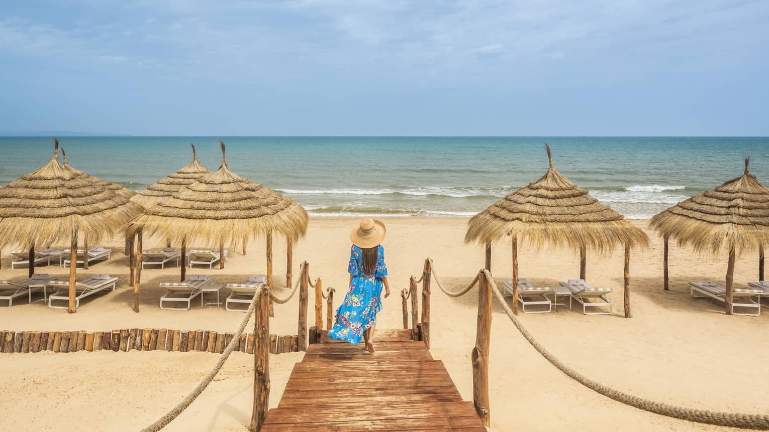 A woman in a floor length blue dress and tan sun hat walks along the wooden boardwalk toward the reed awnings on the beach
