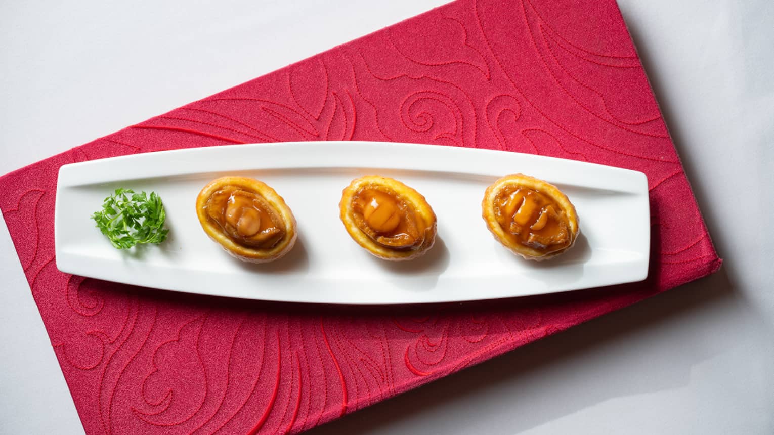 Three small pastries with diced chicken are neatly arranged on a long white plate atop an ornate red cloth. Leafy greens garnish the plate. 