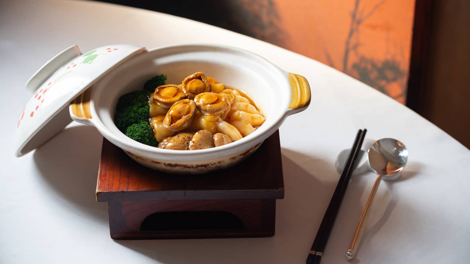 A ceramic dish with a lid on a wooden stand contains a gourmet scallop and green vegetable delicacy. Accompanied by chopsticks and a soup spoon.