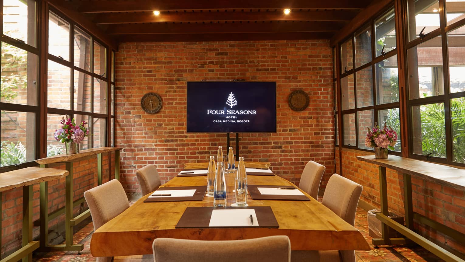 Large rectangular wooden table set for five in a private room with a brick accent wall, two walls of windows and a wood-beamed ceiling