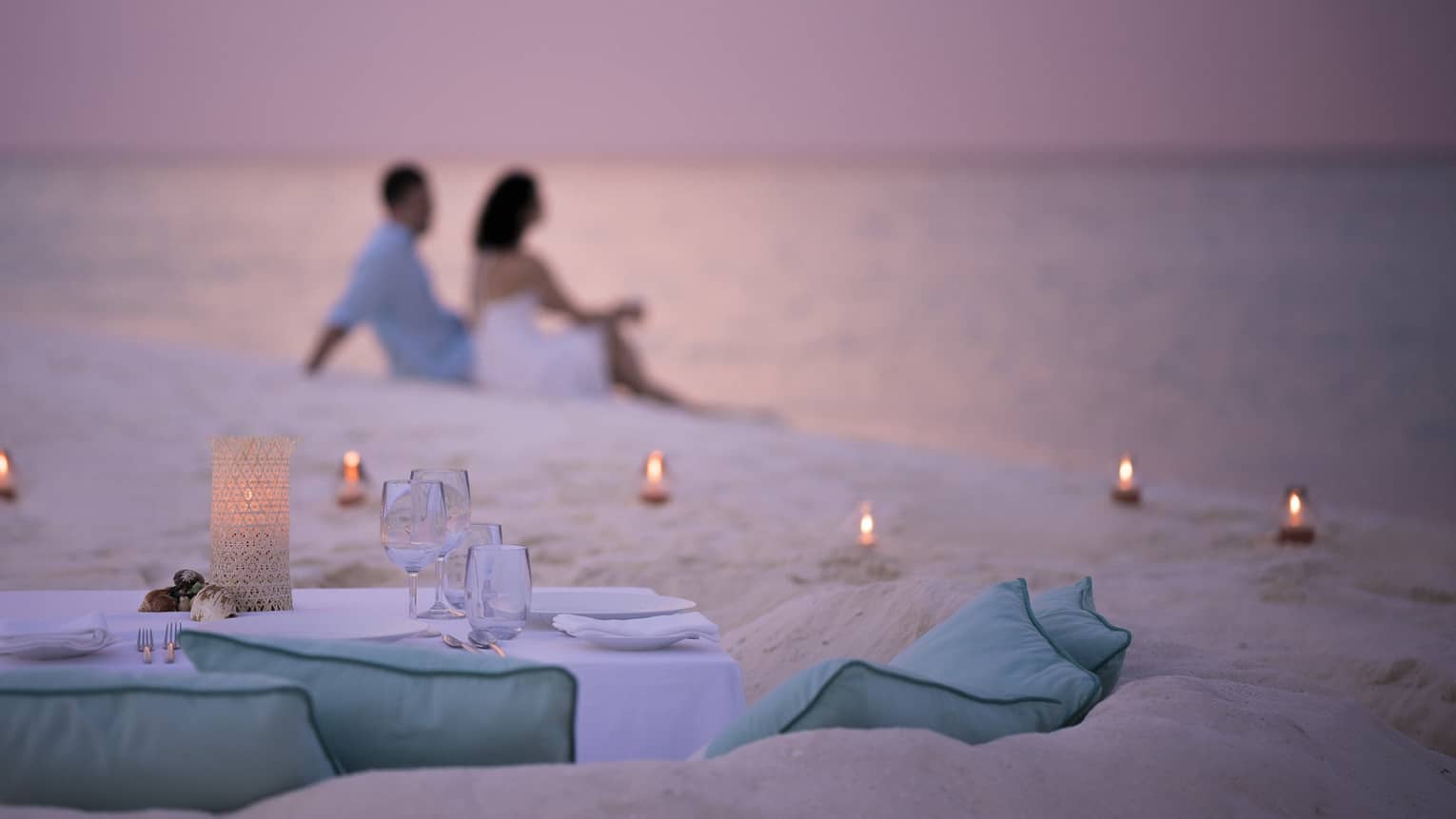 Private dining table in front of sand beach with tealight candles, couple sitting in sand