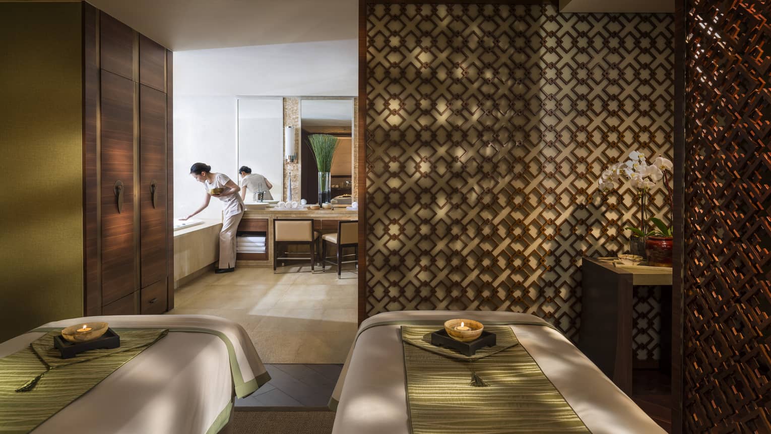 Couples massage beds side-by-side in spa room with candles, wood detail wall, staff preparing bath