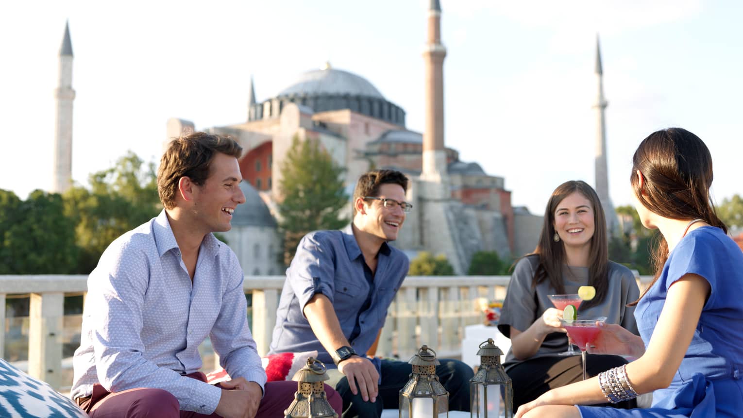 Two men and two women with pink martinis sit on patio in front of balcony, mosque in background