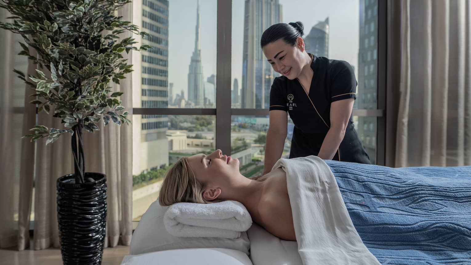 A guest enjoys a massage from a Hotel employee. Behind them, a window frames tall glass skyscrapers and clusters of trees.