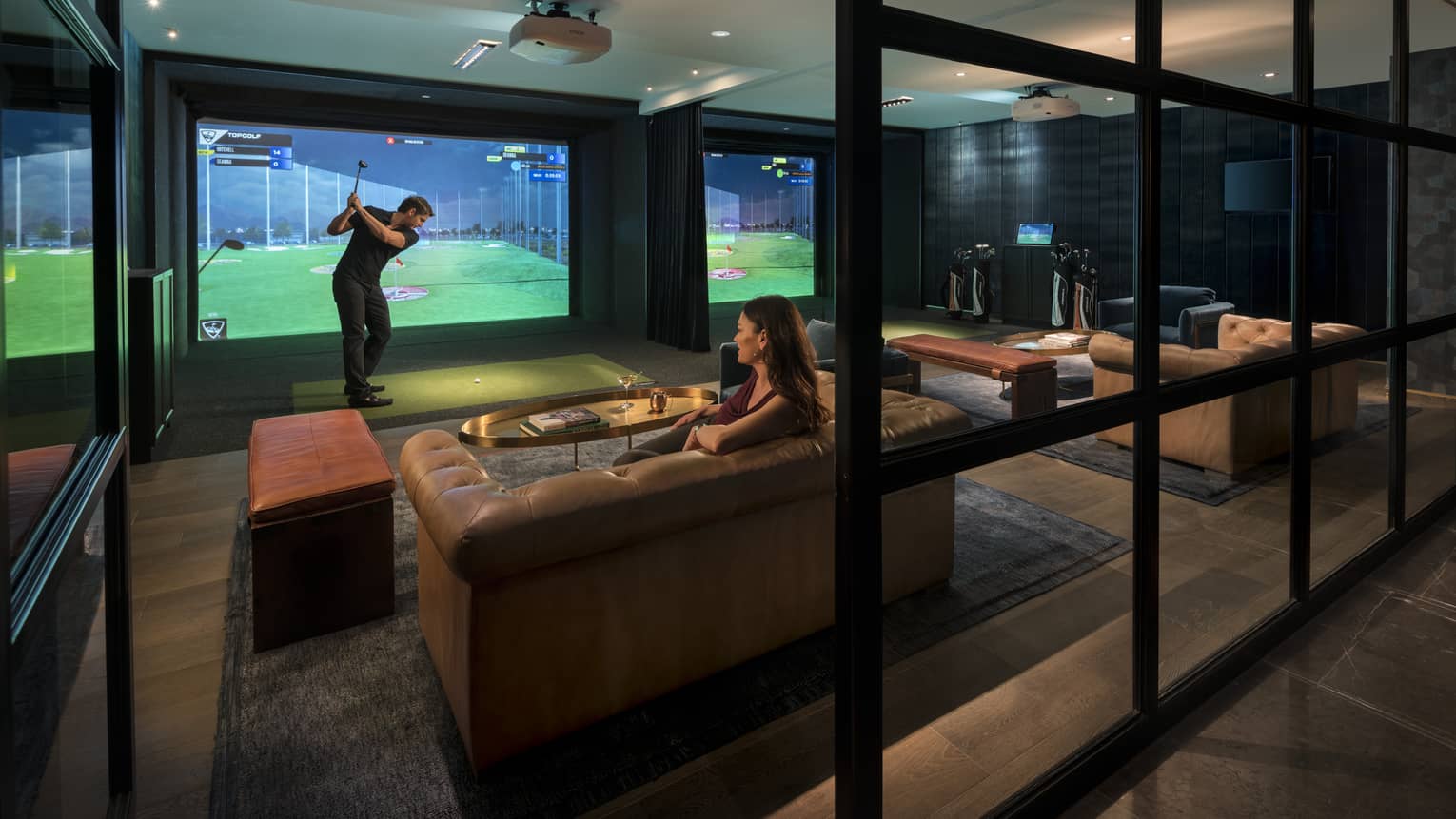 Topgolf lounge, man swings club by large golf simulator screen, woman sits on leather sofa behind him