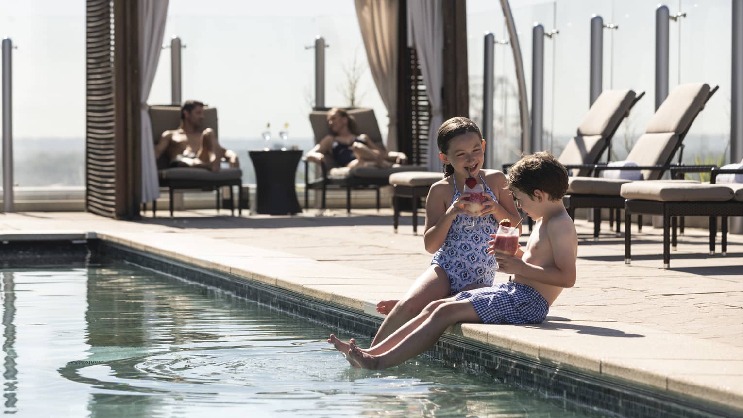 Two children sitting by the edge of a pool, enjoying drinks, while adults relax on lounge chairs under shaded cabanas in the background