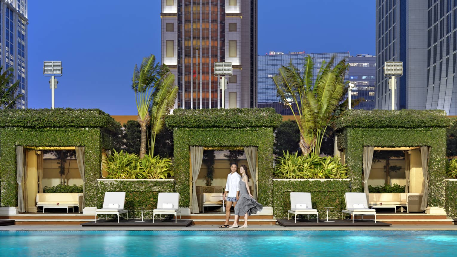 A couple smiles next to a pool, behind them a row of square archway cabanas covered in greenery and flanked by tall palms.