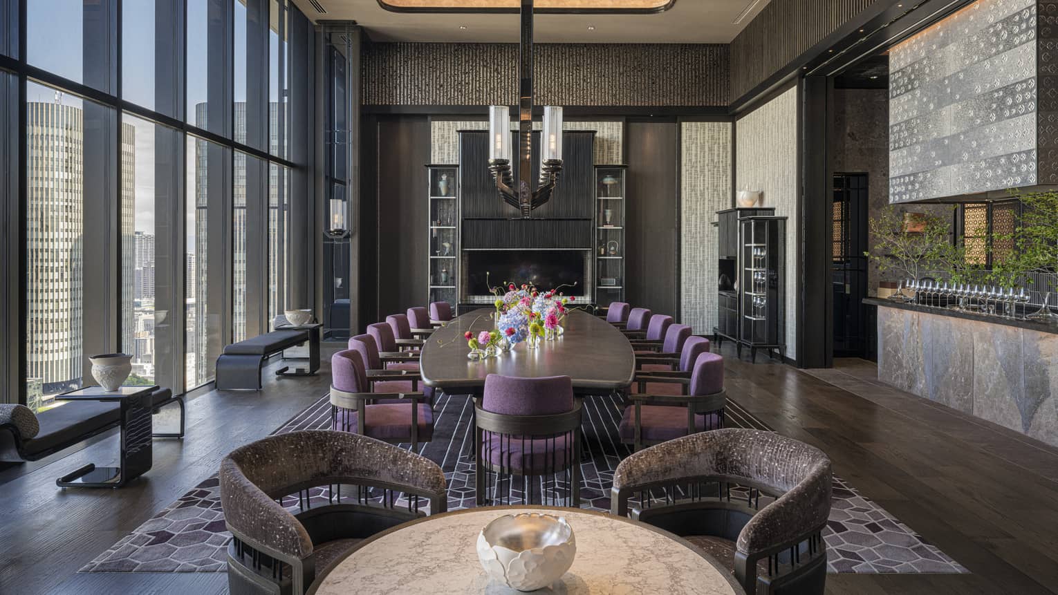 A large well-lit room has dark flooring, an area rug, decorative shelving, accents of greenery, and small tables and benches next to floor-to-ceiling windows along one wall, as well as a round table with chairs and a large oblong table with chairs along its length and topped with an assortment of flowers in vases