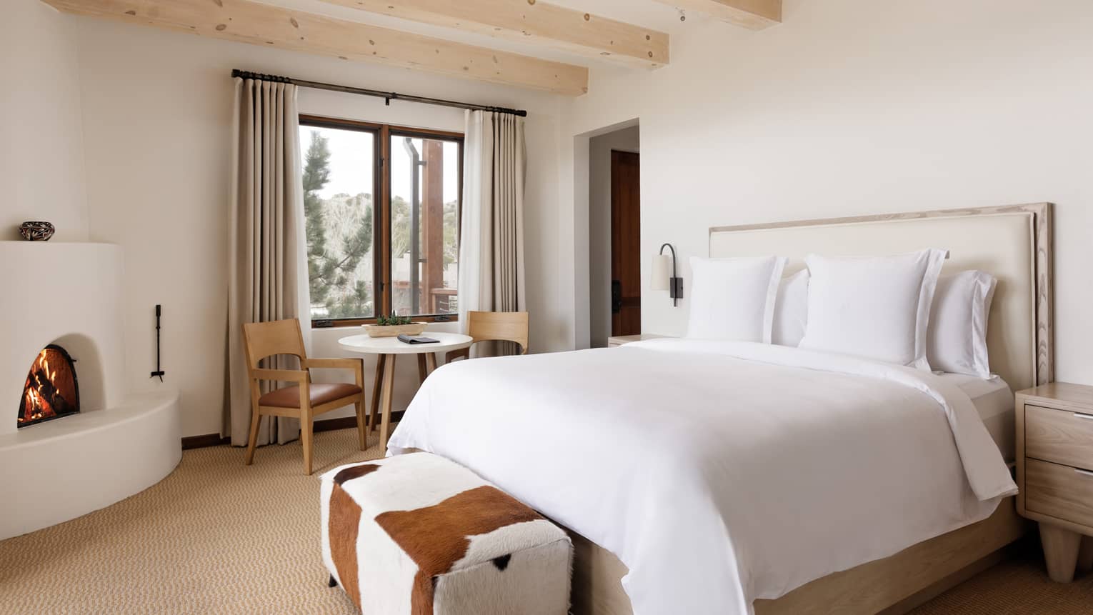 Guest room with king bed, cowhide bench, windowside table and chairs, and beamed ceiling at Four Seasons Resort Santa Fe
