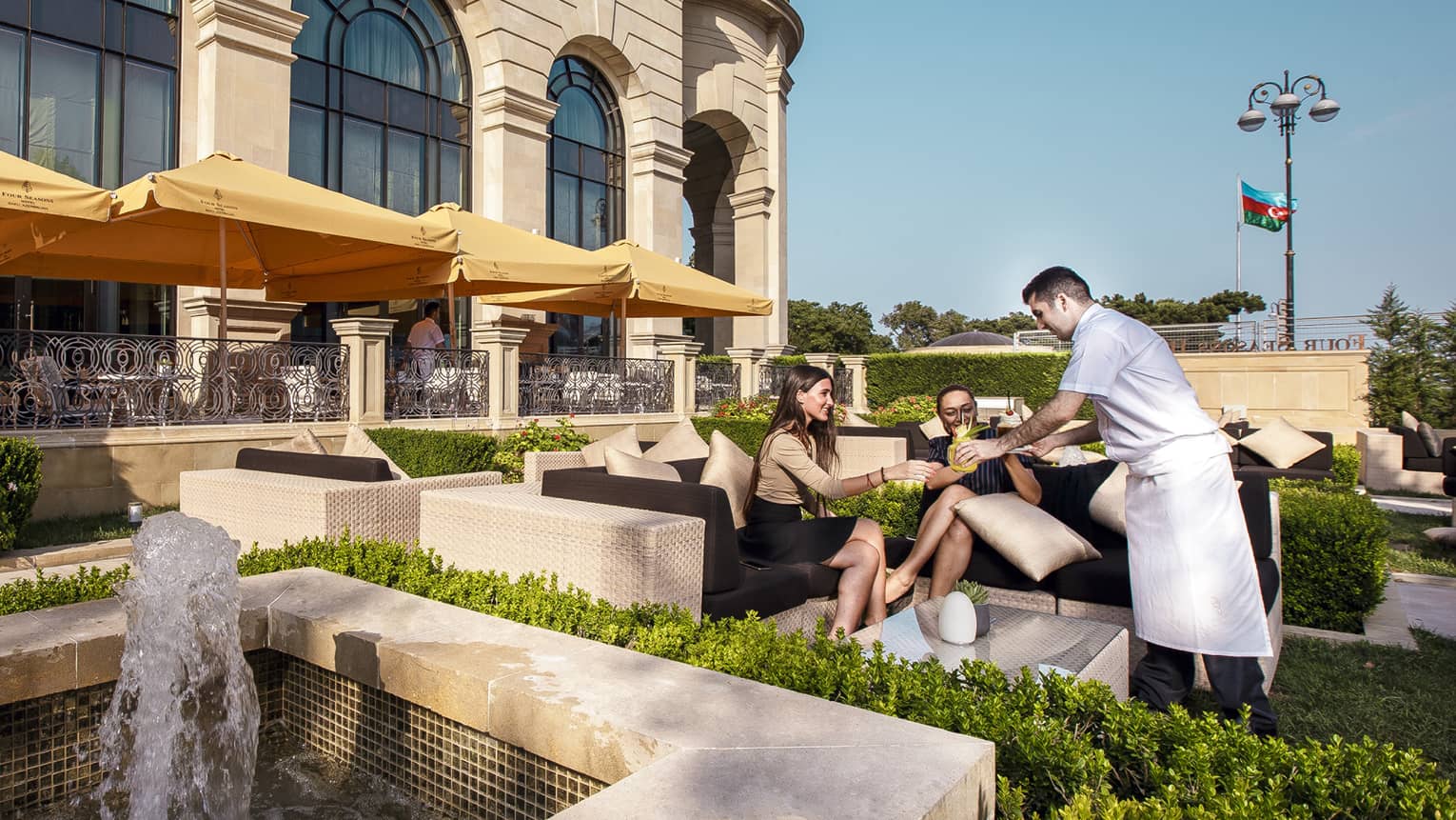 Hotel staff serves cocktails to two women on wicker sofa in large outdoor patio