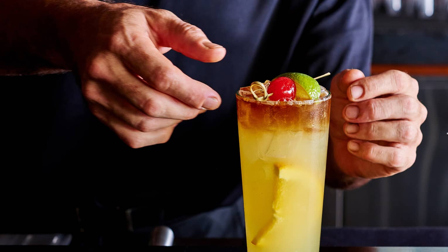 Close-up of hands adjusting a tall orange cocktail on ice, garnished with a cherry and a lime wedge on a small bamboo skewer.