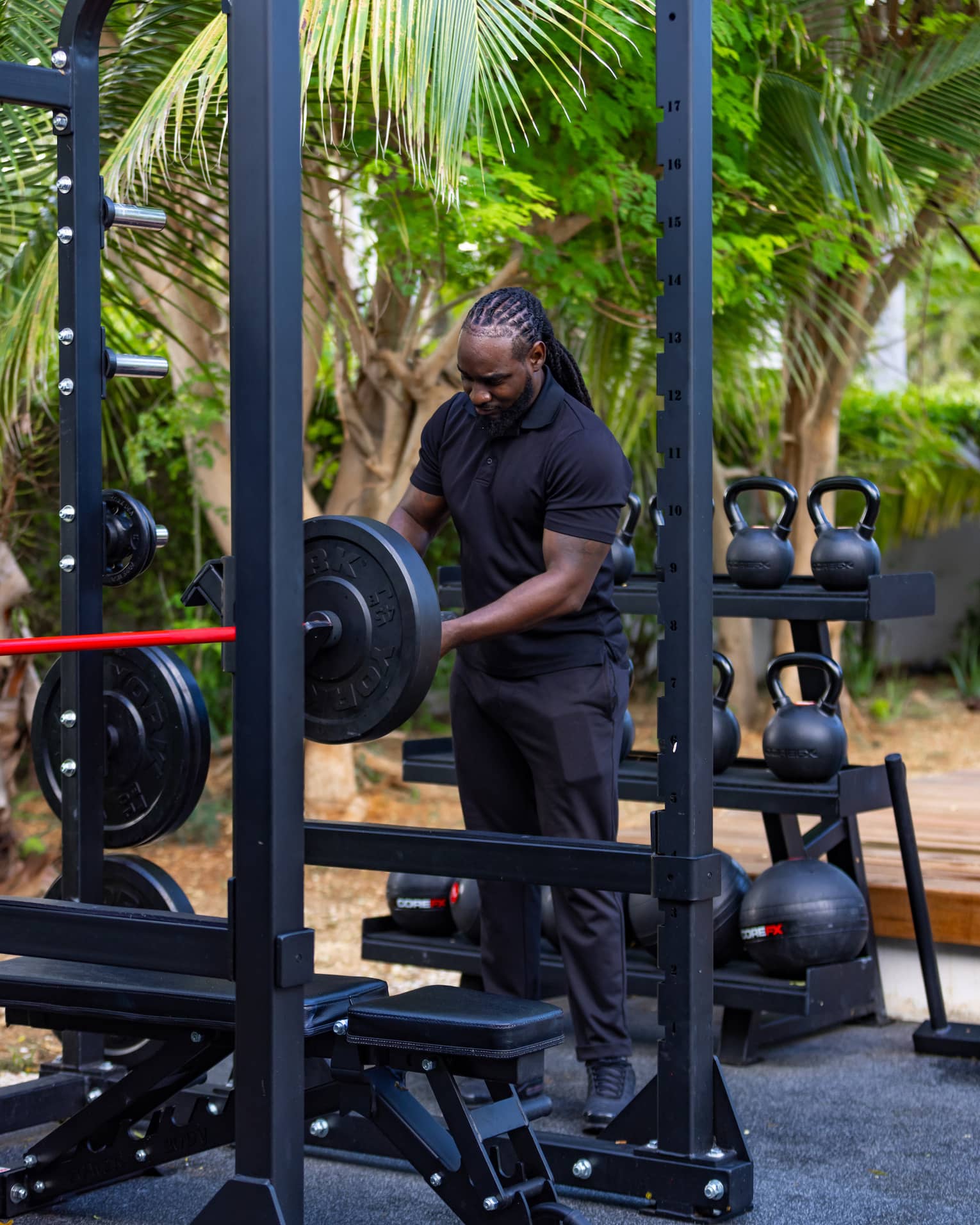 A guest adjusts a barbell in an outdoor gym against a rack of kettlebells and medicine balls and a grove of shady trees.