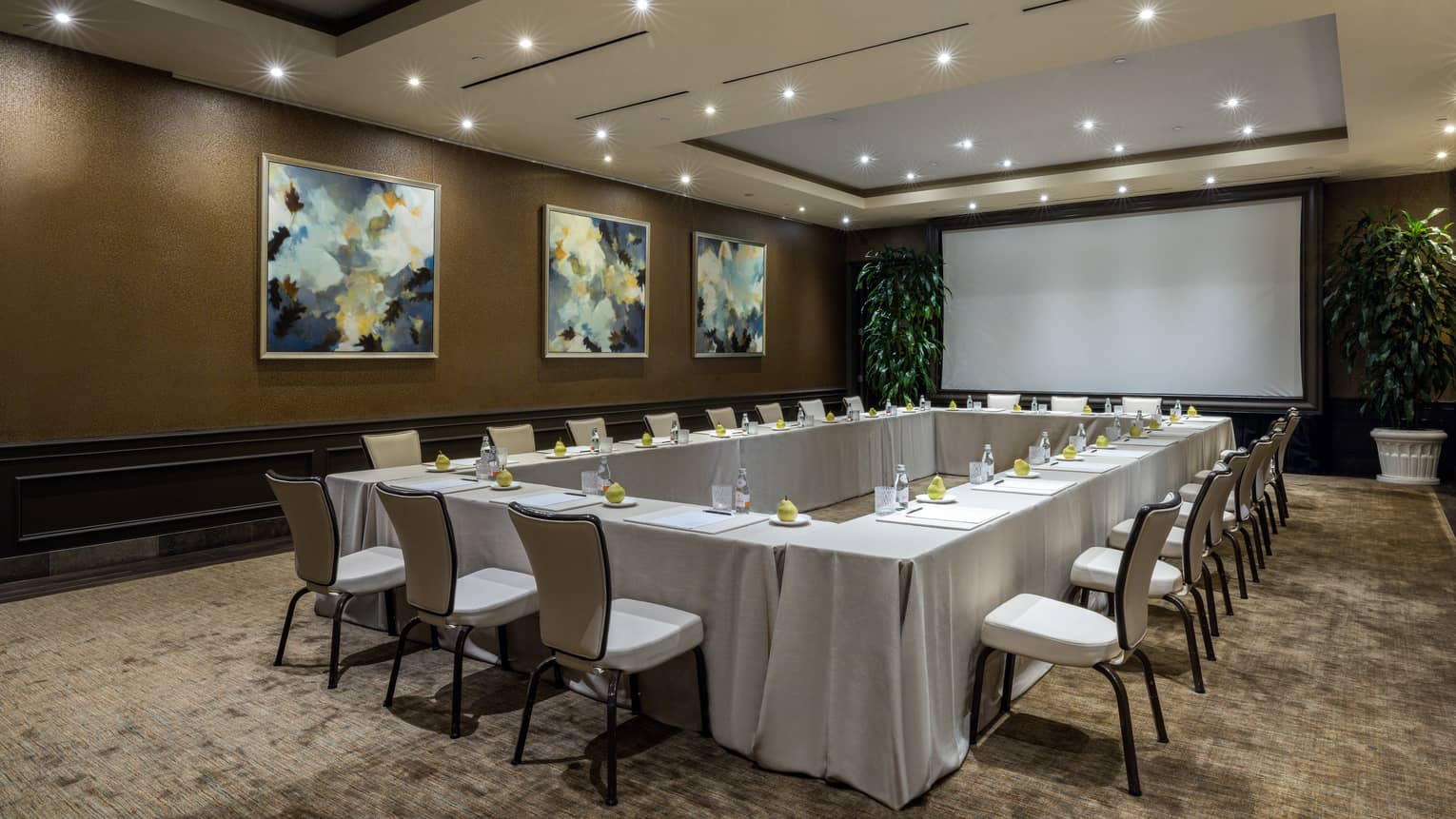 Meeting room set up hollow square with cream-coloured linens, ambient lighting, projection screen framed by large potted plants, and trio of wall paintings