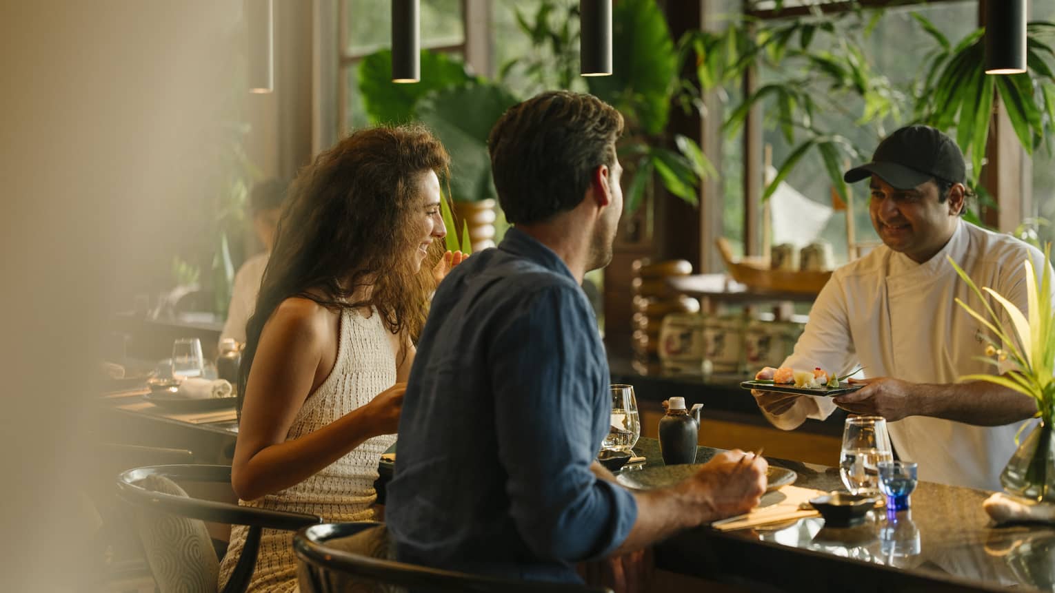 A smiling couple sit at a bar top in a restaurant decorated with vibrant plants as a chef hands them a sushi platter.