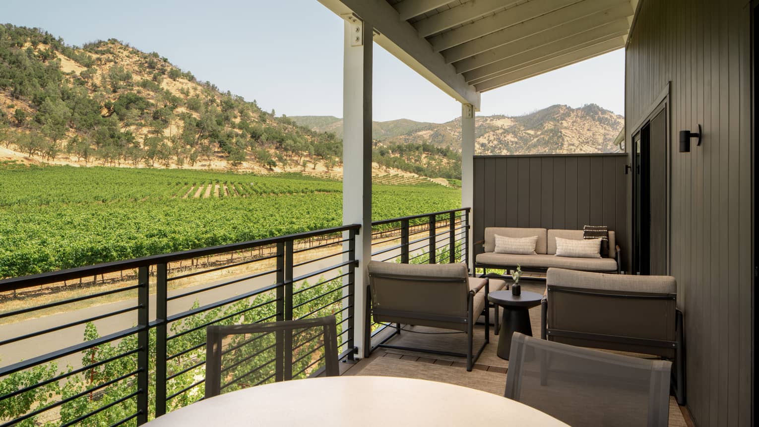 Covered private patio of a resort suite, featuring lounge area and dining table, set next to mountain and lush vineyard