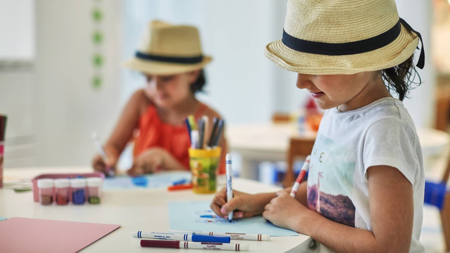 Two smiling children wearing straw fedora hats colour pictures with markers at craft table