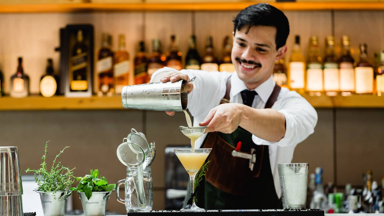 Smiling bartender pours orange cocktail from shaker into martini glass at bar 