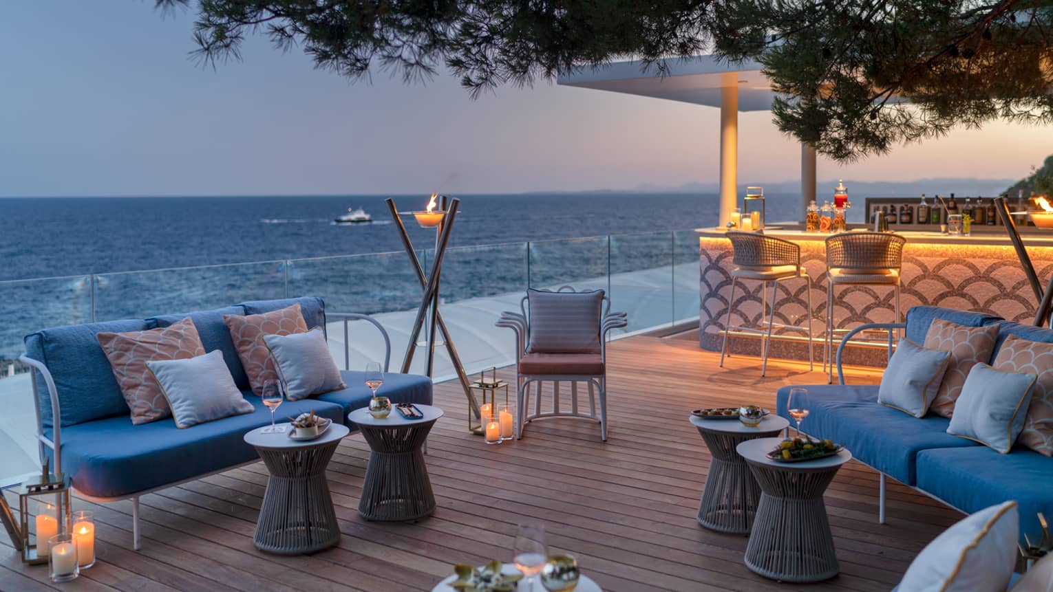 Club Dauphin wooden terrace, blue love seats, orange pillows, candles, torch, uplit bar, sea view at sunset