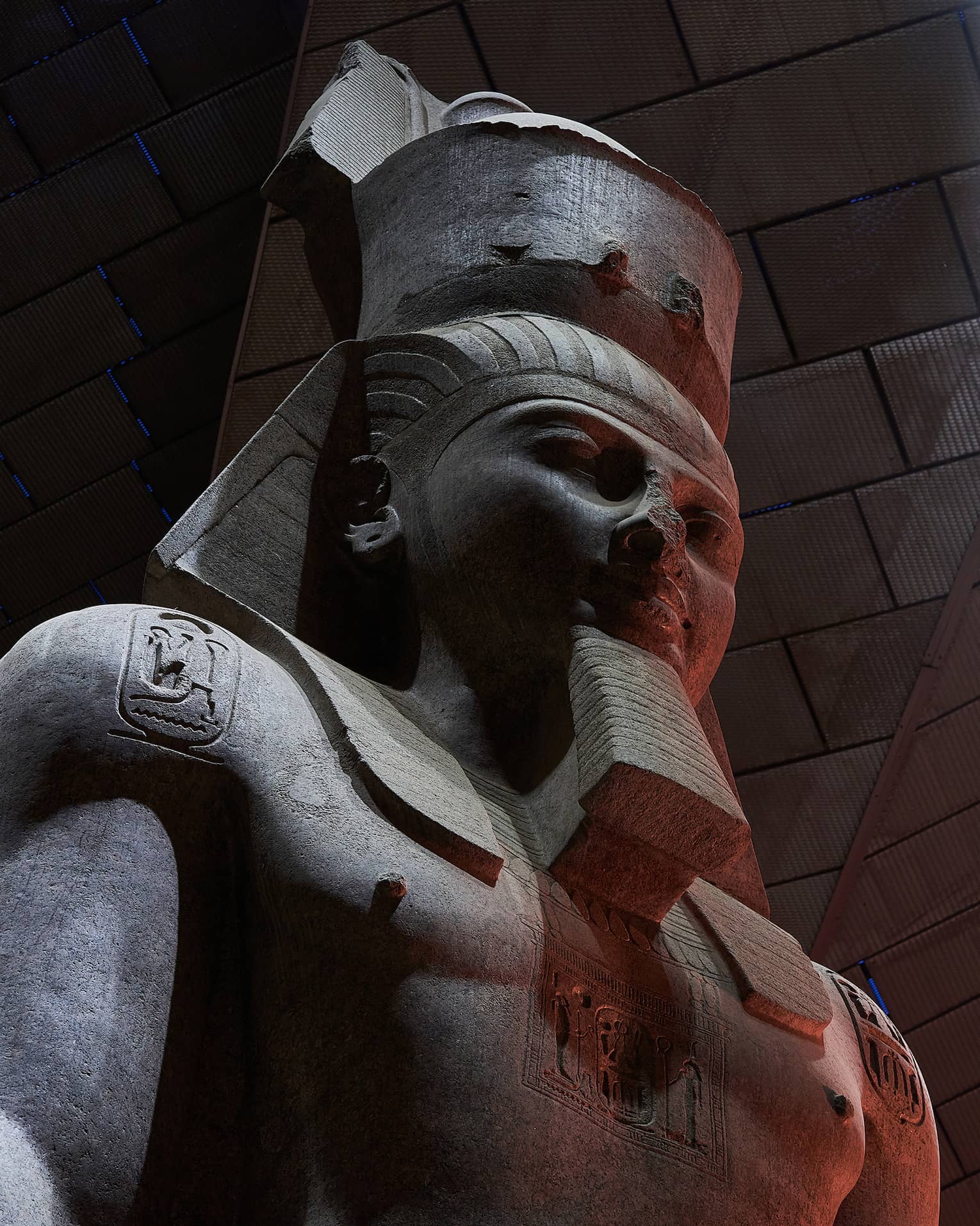 Close-up on a dark stone statue of a bearded pharoah with three heiroglyphic carvings on his chest and shoulders.
