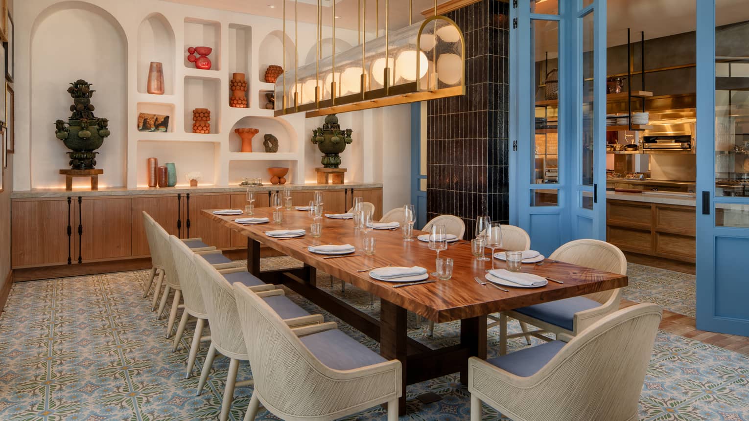 Private dining room with a wood, rectangular table, 11 cream-coloured chairs and a rectangular light fixture overhead, with a wall of arched shelving filled with statues as the backdrop and bright blue doors on the side