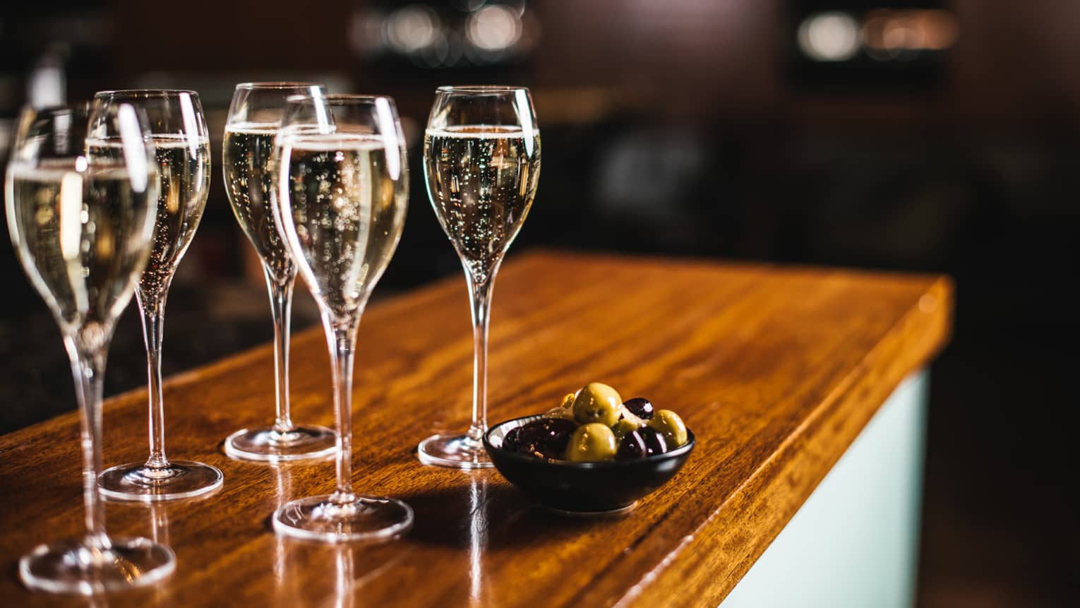 Five glasses of sparkling wine with bowl of black and green olives, atop a wooden bar top