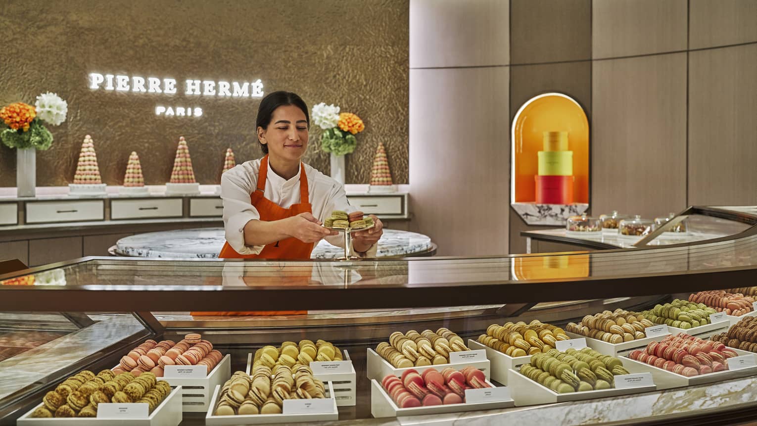 Bakery attendant stands behind a curved glass display case filled with macarons