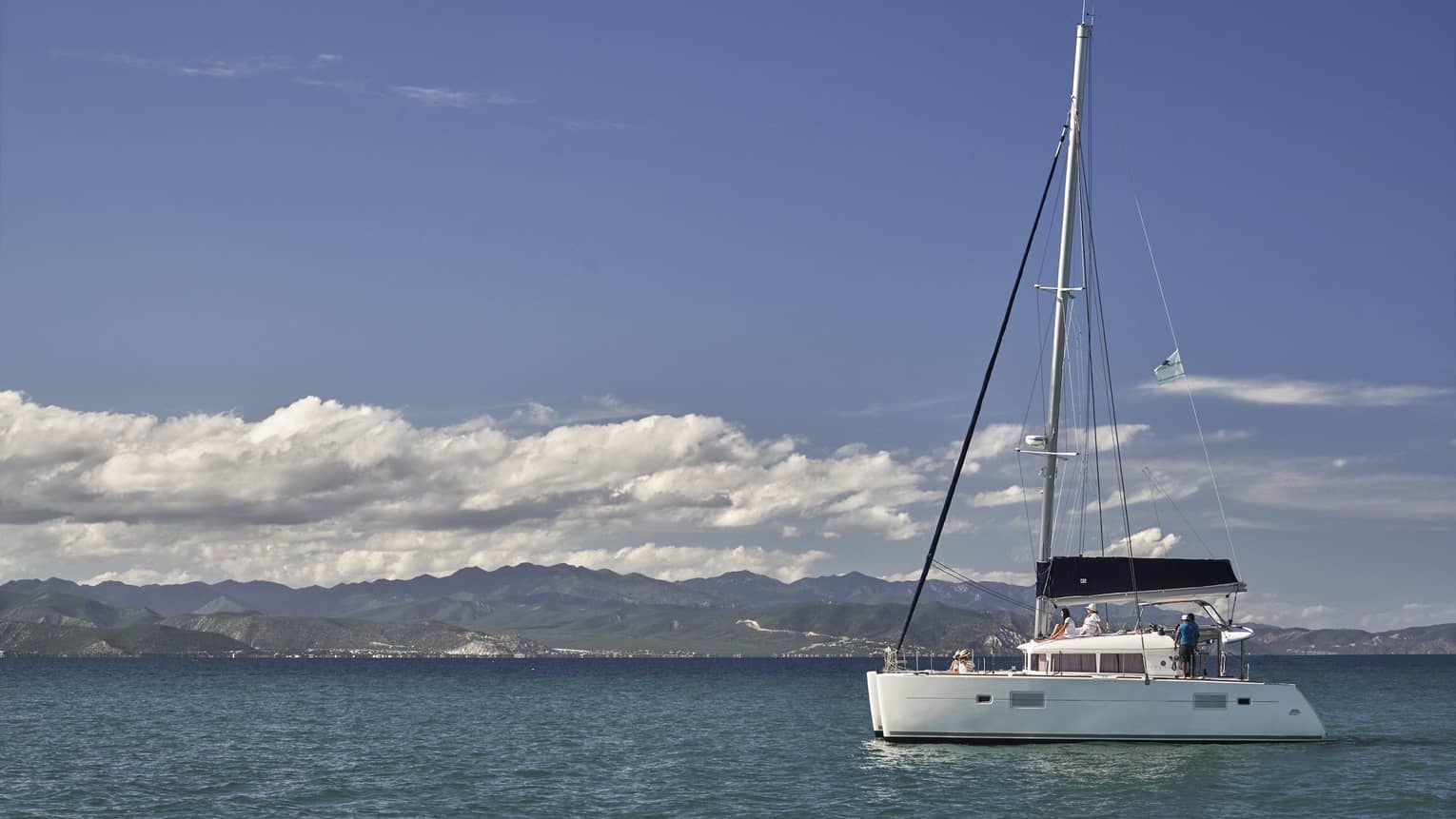 A catamaran on calm, blue water under a partly-cloudy sky with low green hills in the background and mountain ranges beyond.