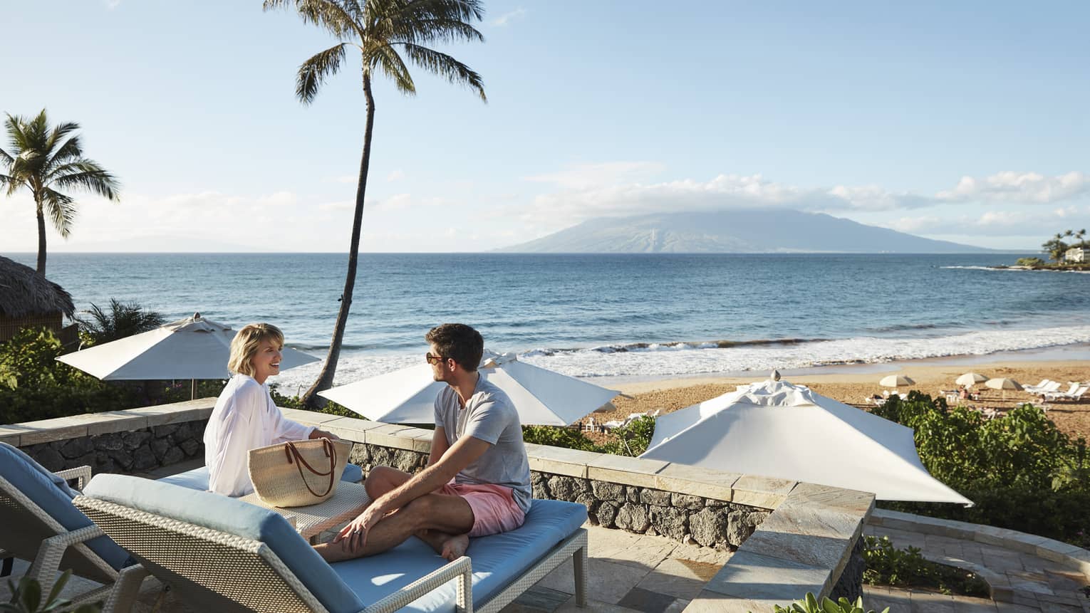 Man in shorts and smiling woman in beach shirt sit on edge of lounge chair, beach and ocean in background