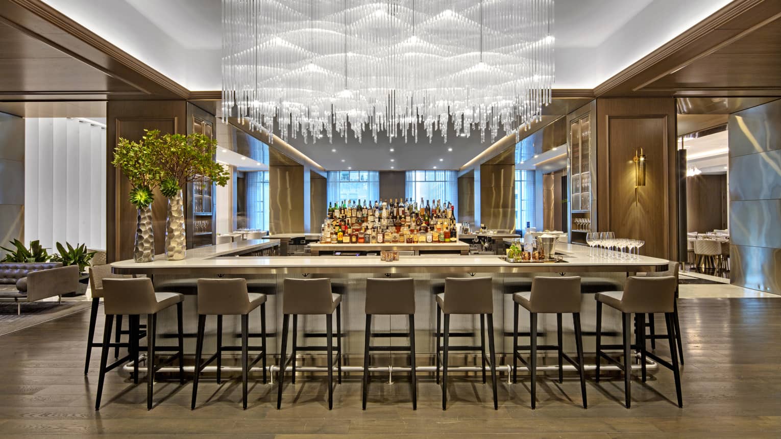 A large bar area with hanging glass chandelier and many grey stools.