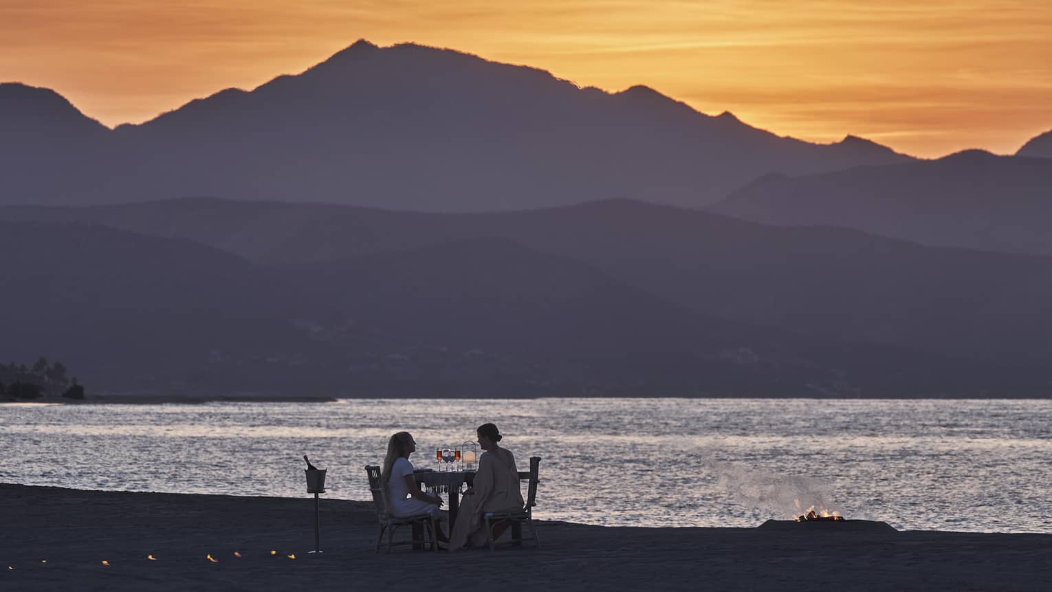 Mountains soar beneath the glow of a bright orange sky. Two people sit at a table by the ocean beside a flickering bonfire.