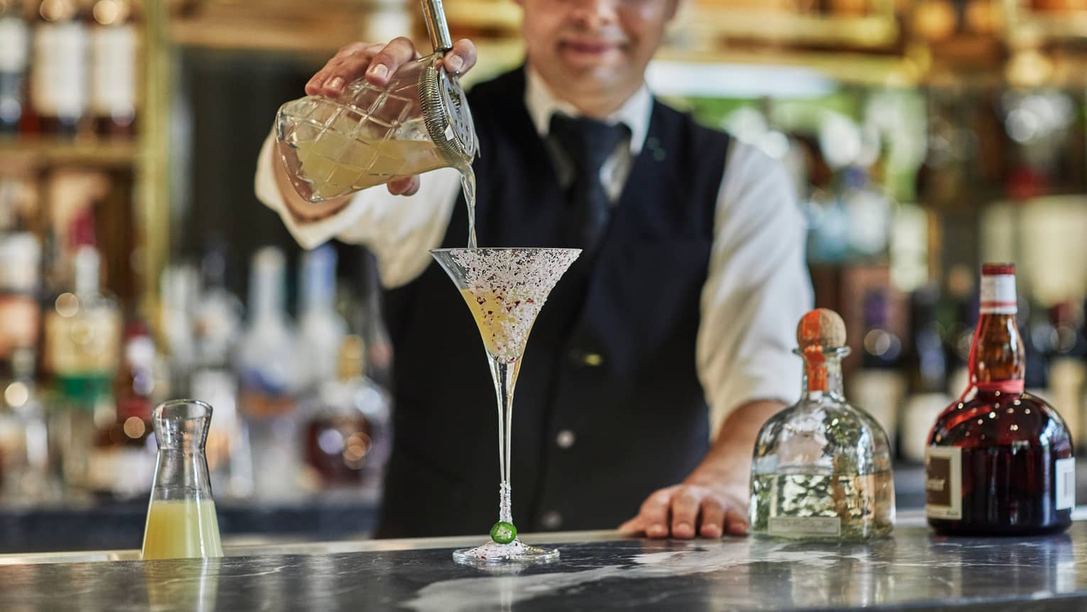 Bartender wearing black vest pours cocktail from crystal shaker into martini glass