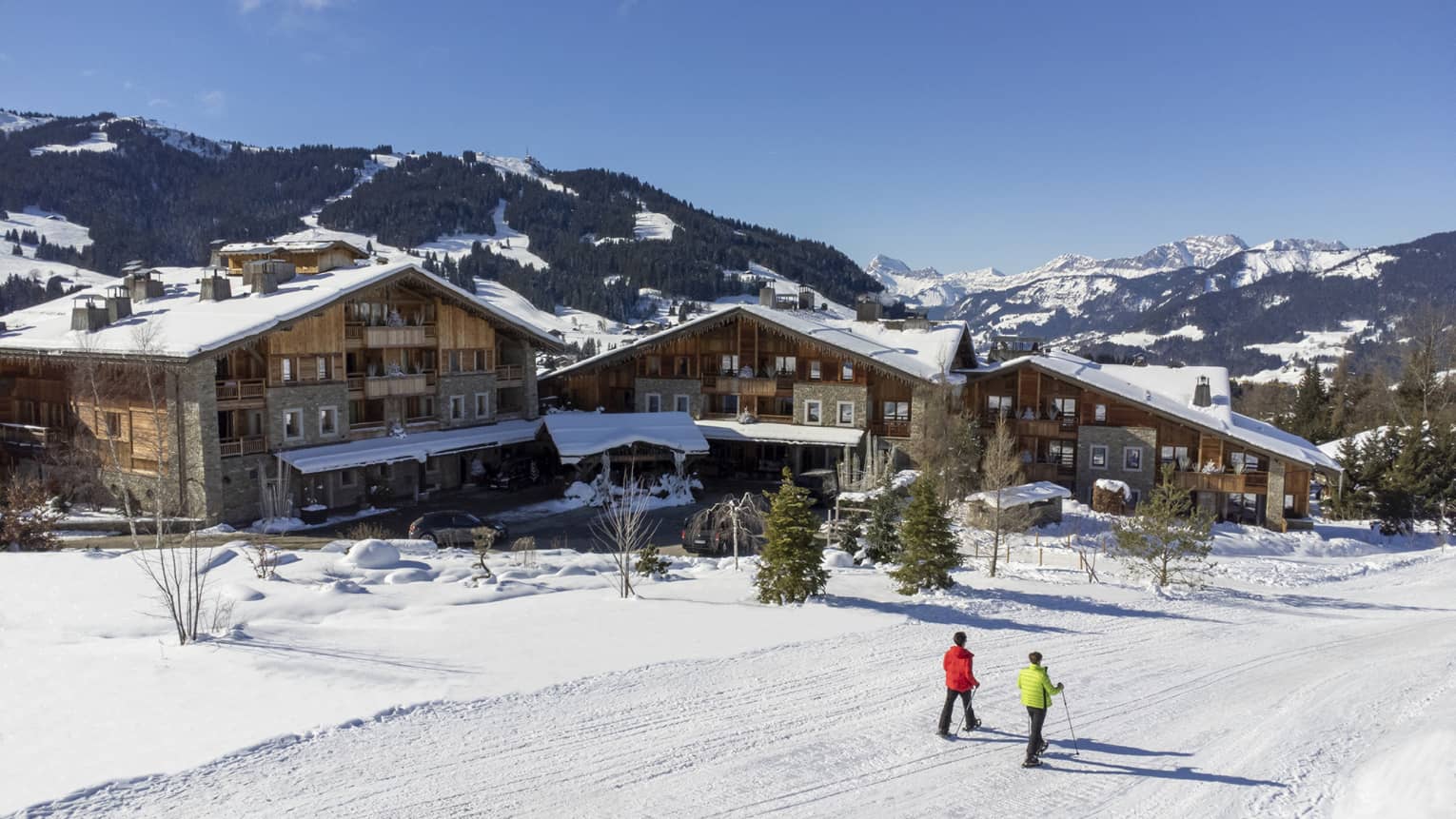 Two people show shoeing on snow-covered path in front of Four Seasons Hotel Megeve with Alps in backdrop