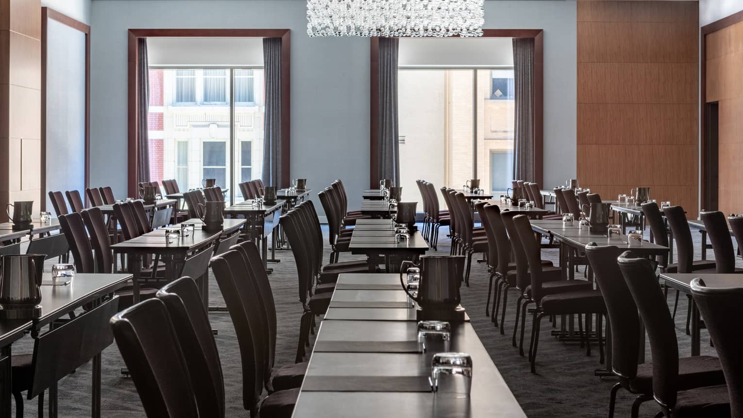 A meeting room with rows of tables and black chairs, there is also a large chandelier and large windows.