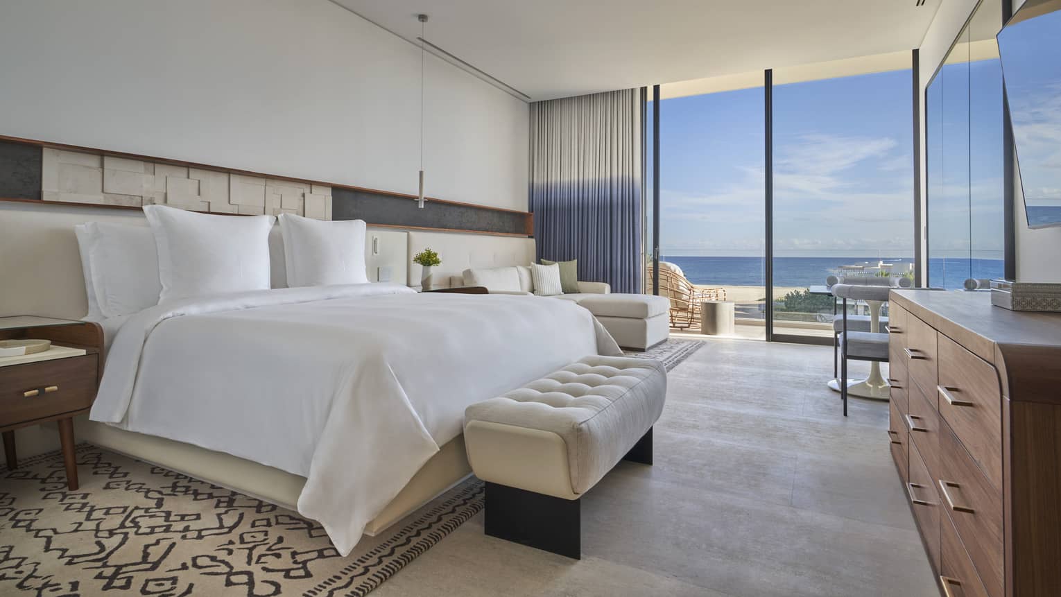White bed with upholstered foot bench, side tables, dresser, lounge chair, tall glass doors and water views