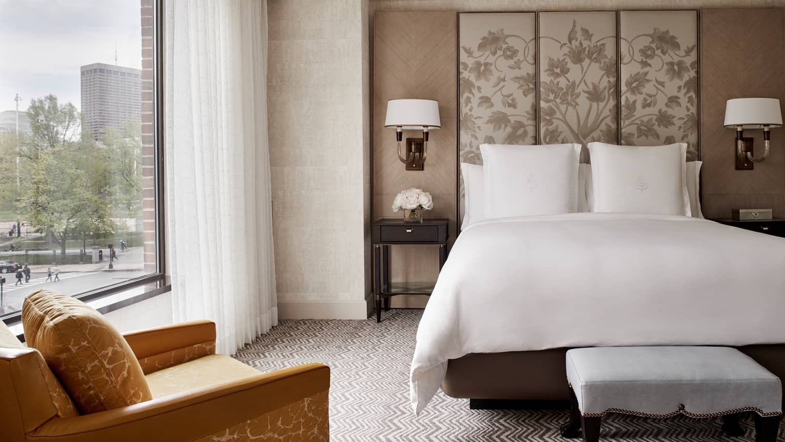 Front view of Executive Suite bed with white linens, beige fabric floral headboard, large window with white curtains, yellow armchair
