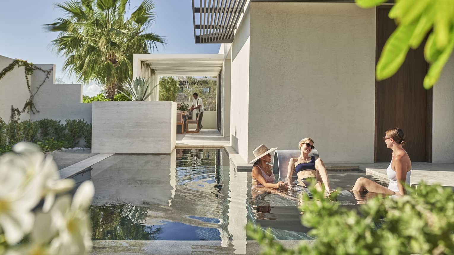 Three people sitting on a shallow pool outside, palm trees and blue skies can be seen behind them.