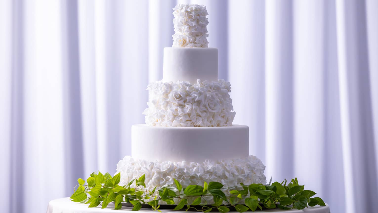 A white floral wedding cake on a stand surrounded by green vines